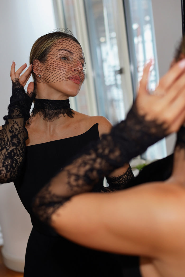 A woman wearing a black lace choker and matching long lace gloves, with a black fishnet veil, posing in front of a mirror in a bright indoor setting.