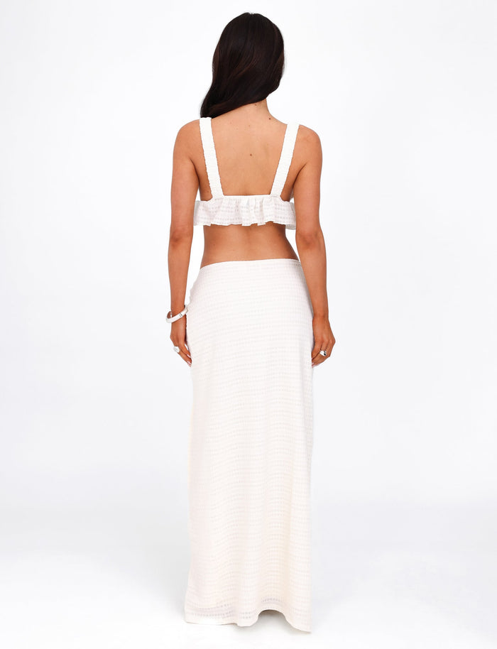 A woman with long dark hair stands facing away, wearing the TIGERMIST ALDRIDGE SKIRT in white, styled with a matching two-piece crochet outfit. The top features wide straps and an exposed back, while the white maxi skirt reaches down to her feet. She accessorizes with a bracelet on her wrist and is photographed against a plain, white background.