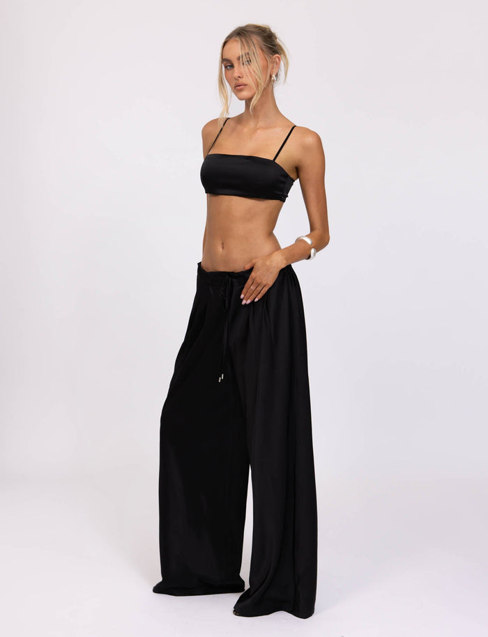 A woman stands against a plain white background wearing the ALINA PANT - BLACK from TIGERMIST. The outfit features a stylish strappy bandeau top and wide-legged, high-waisted pants. Her hair is tied back with loose strands framing her face as she poses with one hand on her hip.