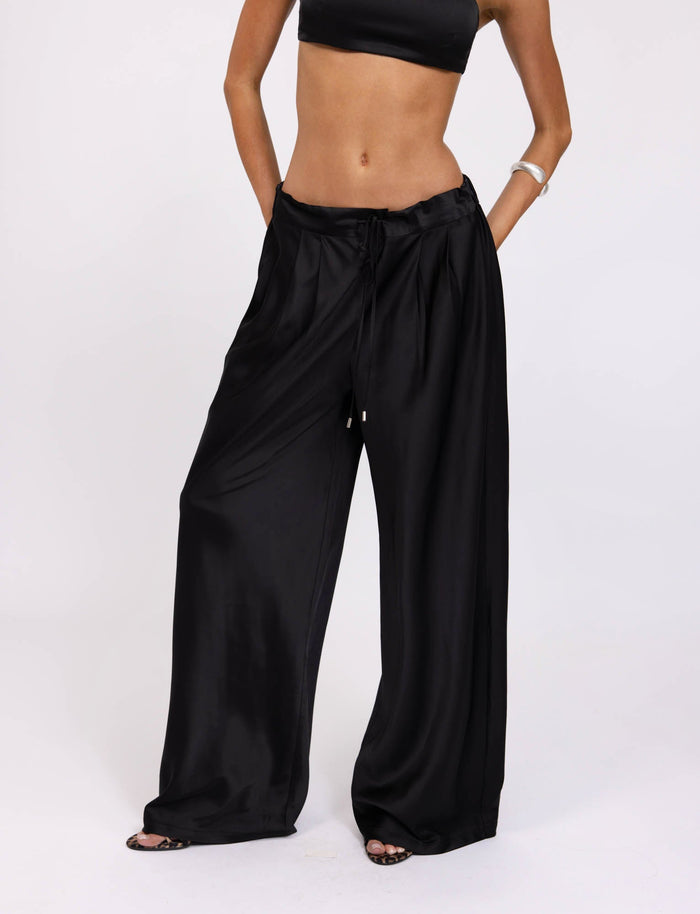 A person is sporting a black crop top and the ALINA PANT - BLACK from TIGERMIST, featuring wide-legged, loose-fitting pants with a drawstring waist. The image highlights the midsection and legs, showcasing the flowy, satin-like fabric of the pants against a plain white background.
