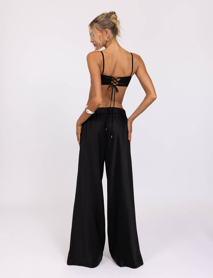 A woman is standing with her back to the camera, dressed in a sleeveless TIGERMIST crop top that ties at the back and the ALINA PANT - BLACK, high-waisted and wide-leg. Her hair is styled in a loose bun, and she poses with one hand on her hip against a plain white background.
