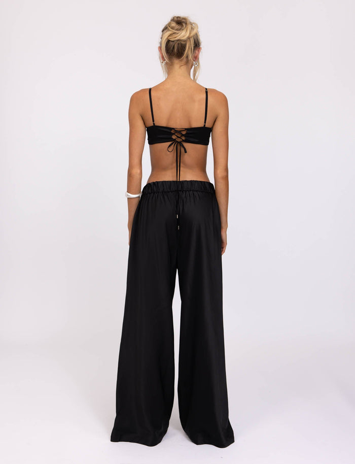 A person with blonde hair tied up is seen from behind, wearing a black Tiger Mist crop top with thin straps and tie detail at the back, paired with the ALINA PANT - BLACK by TIGERMIST. They are standing against a plain white background.