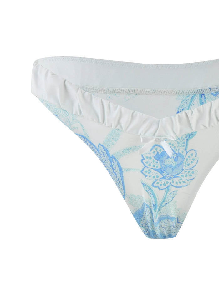 Close-up of the AMARAN BIKINI BOTTOM - BLUE : FLORAL TAPESTRY by TIGERMIST, featuring a cheeky style with a white and light blue floral tapestry pattern. The intricate flowers and leaves provide a delicate, feminine look. The fabric appears smooth, complemented by a slightly ruffled waistband that adds to its charm.