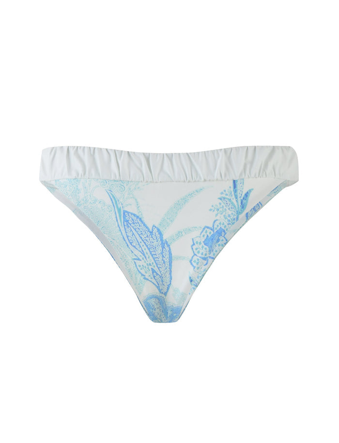 The AMARAN BIKINI BOTTOM - BLUE : FLORAL TAPESTRY by TIGERMIST is a white cheeky bikini bottom adorned with a light blue floral pattern. It features a slightly ruffled waistband and includes stylized leaves and flowers, offering a delicate and summery look.