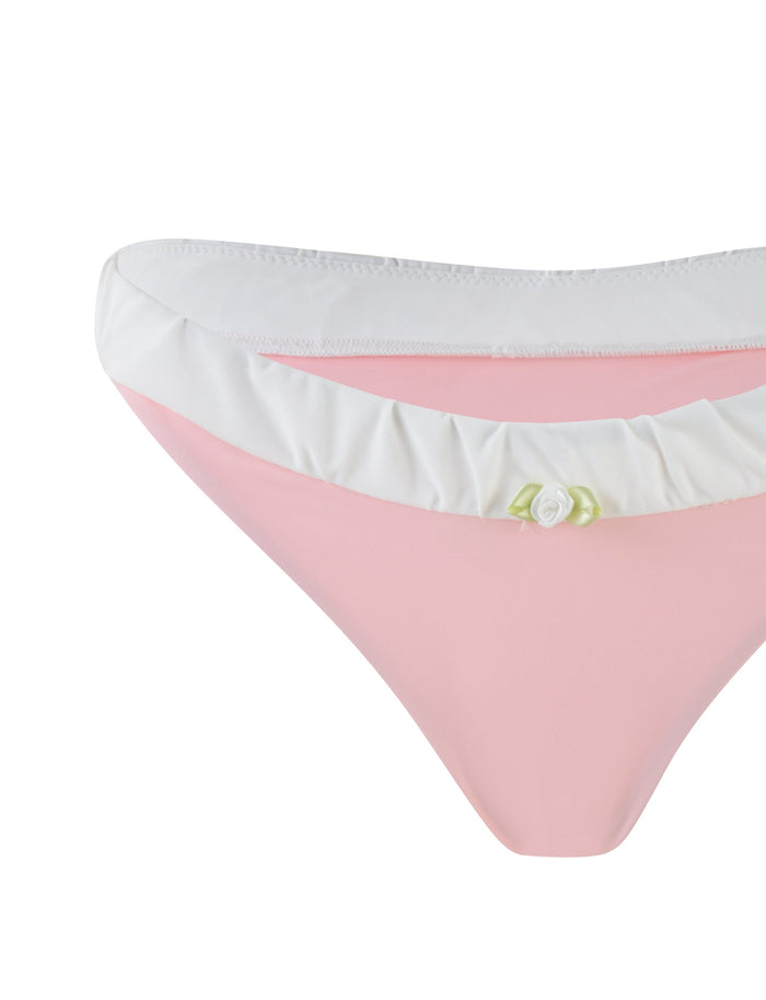 A detailed image featuring the AMARAN BIKINI BOTTOM - PINK from TIGERMIST displays its cheeky cut in pastel pink, adorned with a white ruffle trim along the waistband and accented by a delicate white flower on the front. This image emphasizes the stylish design elements of this swimwear piece.