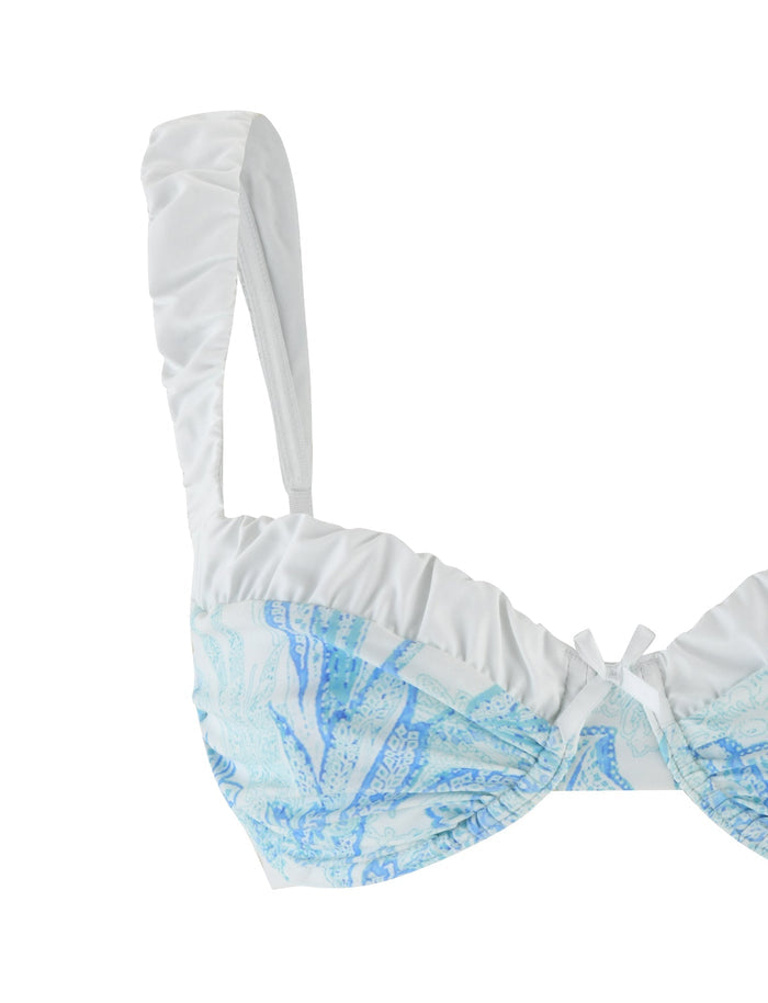 The AMARAN BIKINI TOP - BLUE : FLORAL TAPESTRY by TIGERMIST is showcased in a close-up view, featuring a white balconette design with intricate blue floral and paisley patterns. It includes adjustable straps, ruched detailing at the front, and a small bow accent at the center. The plain white background emphasizes its flattering bodycon fit style.