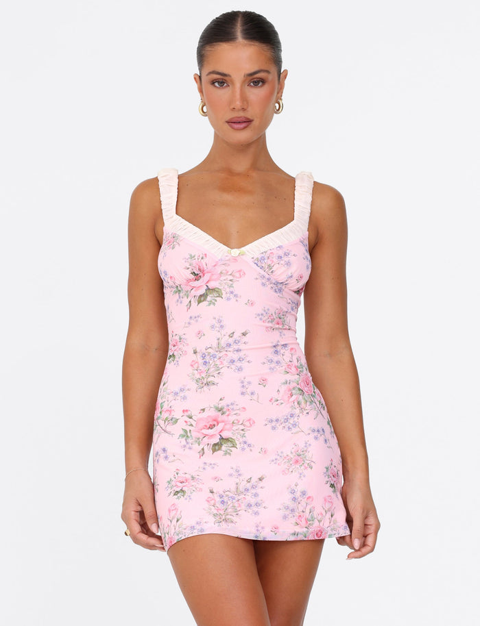 Someone wearing the Amaran Mini Dress by Tigermist, a pink mini dress with a wild flower floral print, poses against a plain background. The dress showcases a V-neckline and ruffled straps. The individual has long hair tied back and is accessorized with earrings.
