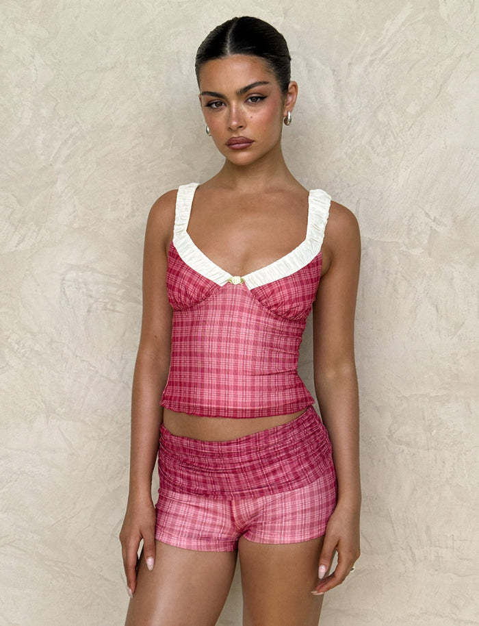 A person stands against a textured beige wall wearing a TIGERMIST AMARAN TOP - RED CHECK, featuring a white ruffled neck detail. They have slicked-back hair, hoop earrings, and a calm expression.