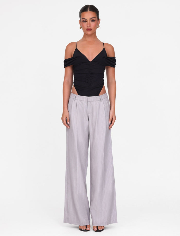 A woman faces forward in a black off-shoulder top with spaghetti straps and TIGERMIST AMBERLY SUIT PANT in grey pinstripe. Her hair is tied back, and she wears open-toe heels against a plain white background.