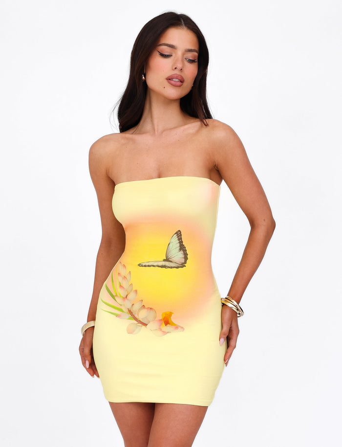 A woman in the AMBROSE DRESS - YELLOW : BUTTER from TIGERMIST, a strapless, form-fitting yellow ombre butterfly dress featuring a detailed flower design at the front, stands against a plain white background. She has long dark hair and is accessorized with gold hoop earrings and bracelets, posing with one hand on her hip.