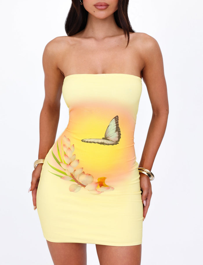 A woman in the AMBROSE DRESS - YELLOW: BUTTER by TIGERMIST, featuring a strapless yellow ombre design with a butterfly and flower print, poses gracefully. She is adorned with gold hoop earrings and bangles on both wrists, her long, dark hair flowing. The bodycon silhouette hugs her figure perfectly as she stands against a plain white background.