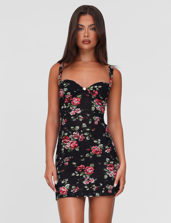 A woman with long brown hair wears the TIGERMIST ANDEE MINI DRESS in BlackRose floral, featuring a fitted, sleeveless lace design with red and pink rose prints, standing against a plain white background.