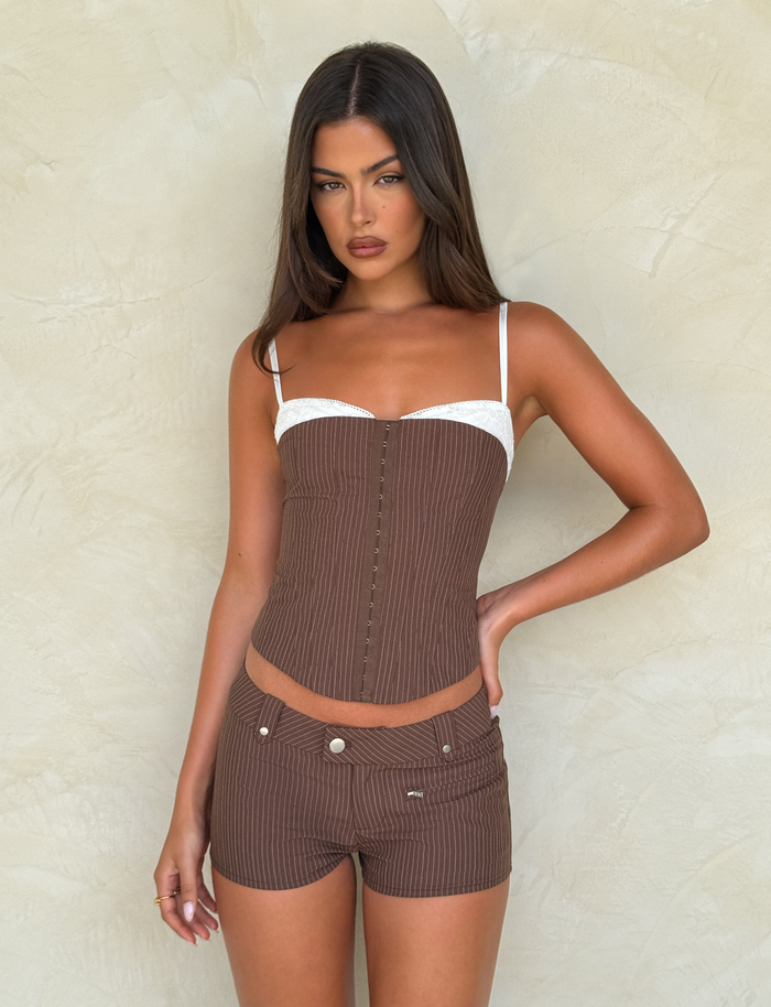 A woman with long brown hair wears the TIGERMIST ANTONELLA CORSET 2.0 in chocolate pinstripe with adjustable straps and matching shorts, standing against a beige textured wall.