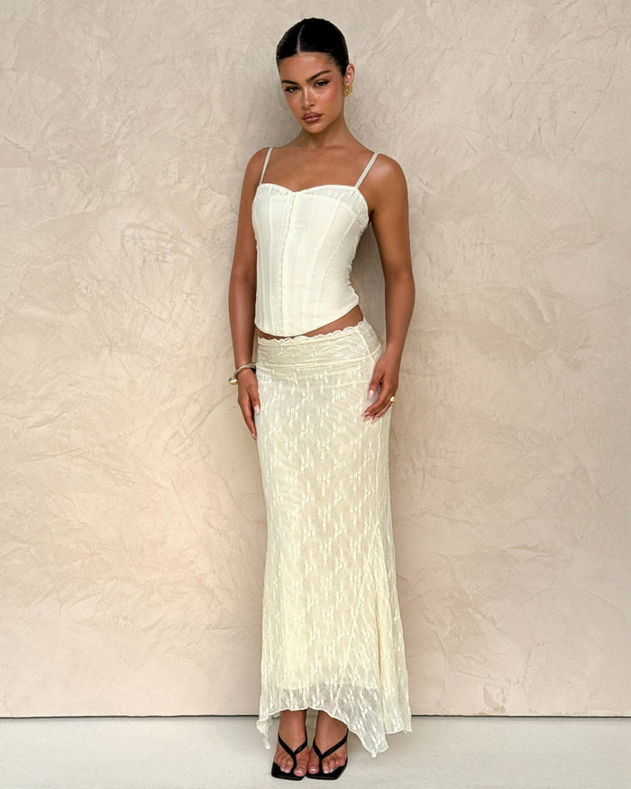 A woman poses against a textured beige wall, wearing the TIGERMIST ANTONELLA MIDI SKIRT in cream lace and a matching cream corset top with thin straps. Her hair is pulled back, and she pairs the look with black sandals.
