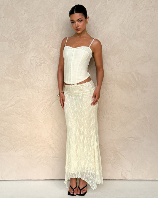 A woman poses against a textured beige wall, wearing the TIGERMIST ANTONELLA MIDI SKIRT in cream lace and a matching cream corset top with thin straps. Her hair is pulled back, and she pairs the look with black sandals.