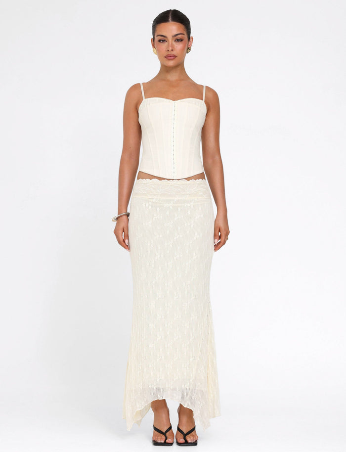A woman stands against a plain white background, wearing the TIGERMIST ANTONELLA MIDI SKIRT in lace cream with a handkerchief hem and a cream corset top. Her hair is pulled back as she looks directly at the camera.