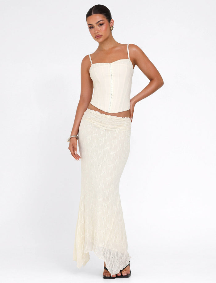 A woman poses against a white background wearing the TIGERMIST ANTONELLA MIDI SKIRT in cream lace, paired with a matching corset top, black sandals, and accessorized with earrings, a bracelet, and her hair pulled back.