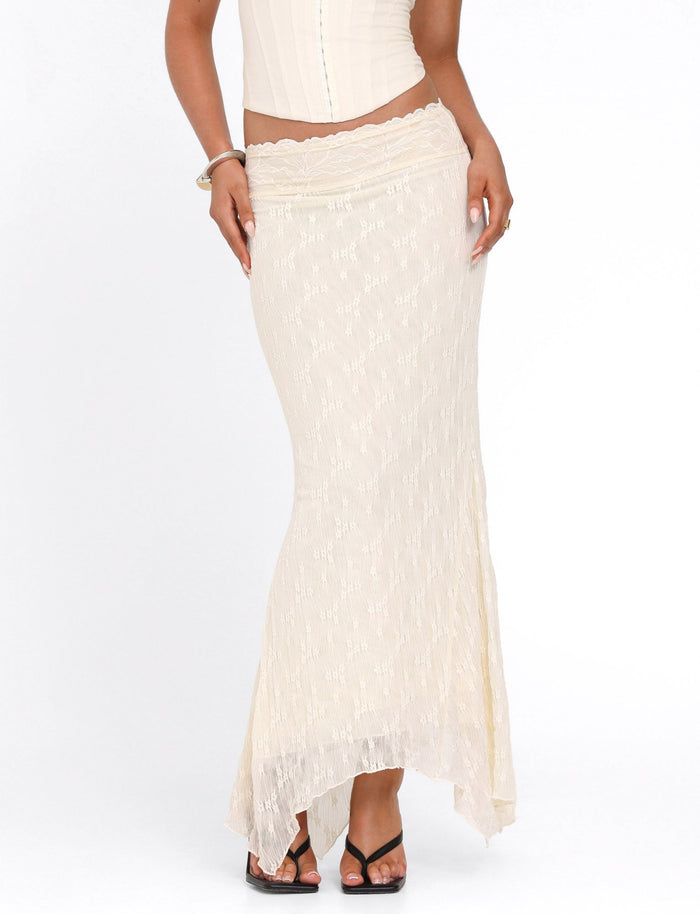 A person wears the TIGERMIST ANTONELLA MIDI SKIRT in cream lace with an uneven hem, paired with a fitted sleeveless top and black heeled sandals, standing against a white background.