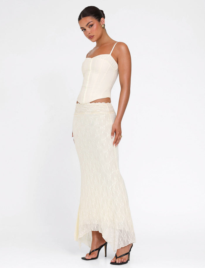 A woman poses against a white background wearing the TIGERMIST ANTONELLA MIDI SKIRT in cream lace, paired with a cream corset top and black high-heeled sandals.