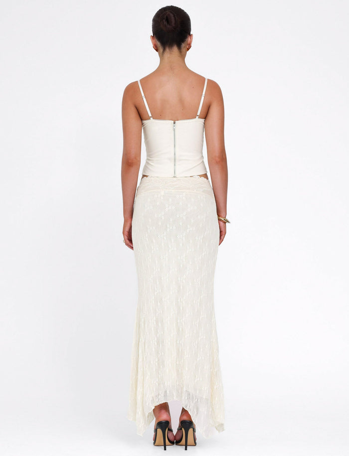A woman with her hair in a neat bun wears the TIGERMIST ANTONELLA MIDI SKIRT in cream lace, styled with a sleeveless top and heels, standing against a plain white background.
