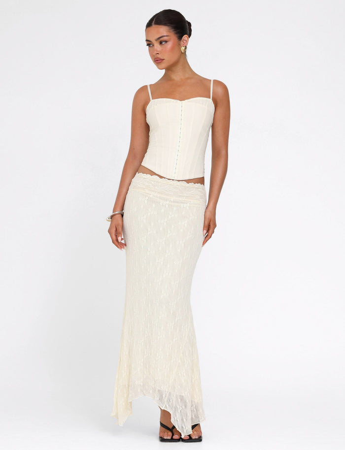 A woman stands against a white background wearing the TIGERMIST ANTONELLA TOP in cream with adjustable straps and a matching long skirt, her hair in an updo and black sandals on her feet, looking to the side.