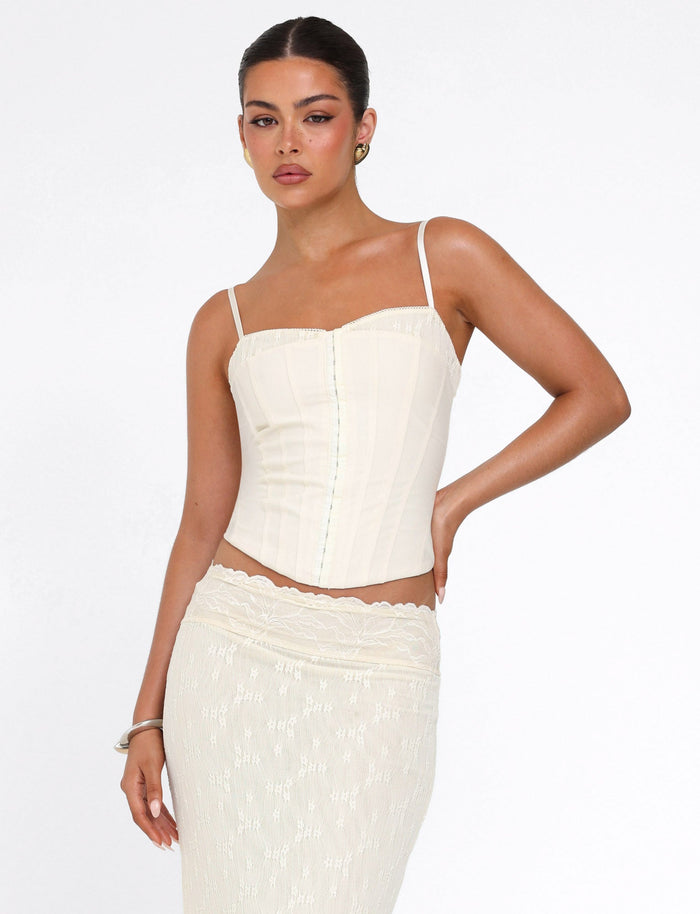 A woman poses against a white background wearing the TIGERMIST ANTONELLA TOP in cream, featuring adjustable straps, paired with a matching lace-patterned skirt. Her hair is pulled back and she accessorizes with earrings and a bracelet.