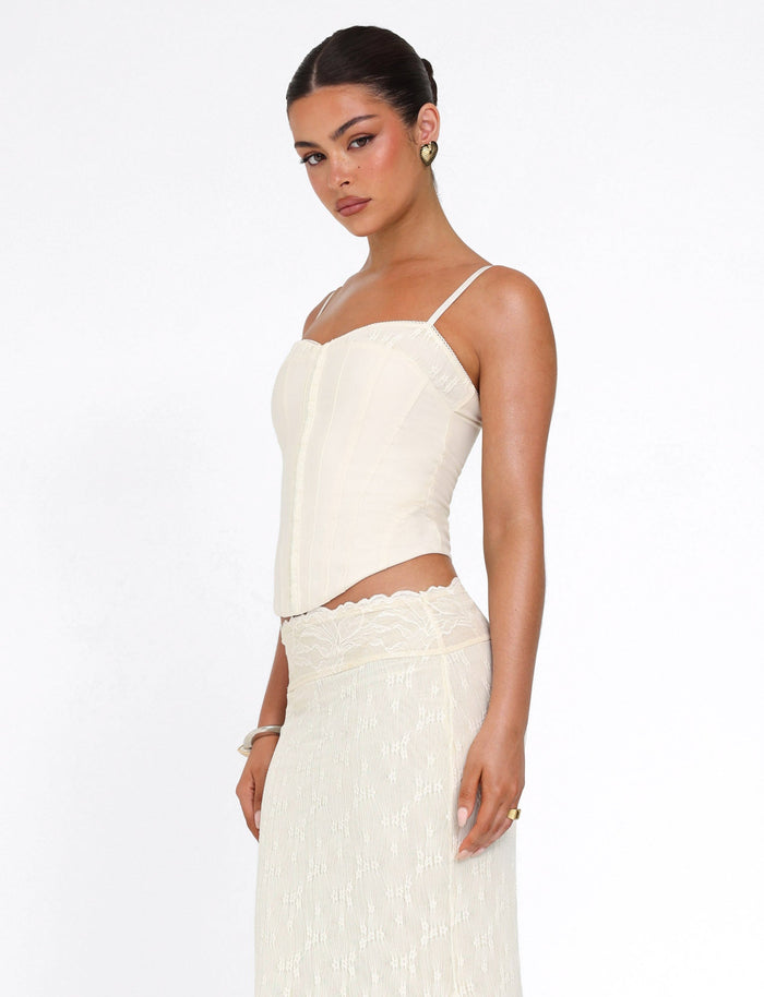 A woman wears the TIGERMIST ANTONELLA TOP in white/cream with adjustable straps and a matching lace skirt, standing against a plain white background and gazing over her shoulder with a neutral expression.