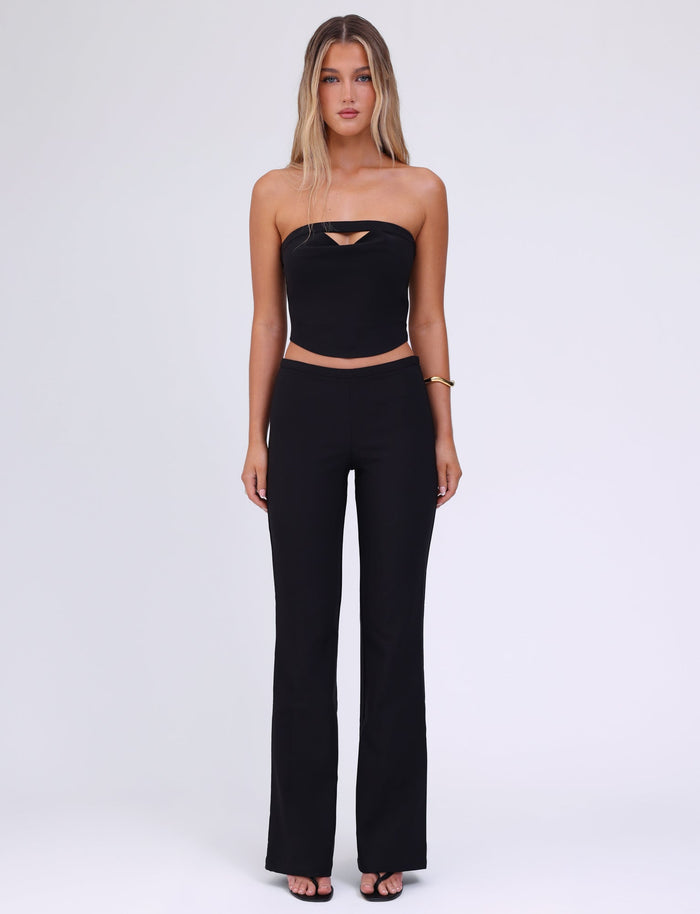 A woman with long, blonde hair stands against a plain white background. She is wearing a black tube top with a small cut-out at the chest, the TIGERMIST AUDRIE PANT in black high-waisted bell-bottom style made from suiting fabrication, and a faux leather belt. She has a gold bracelet on her left wrist and wears black open-toe shoes.
