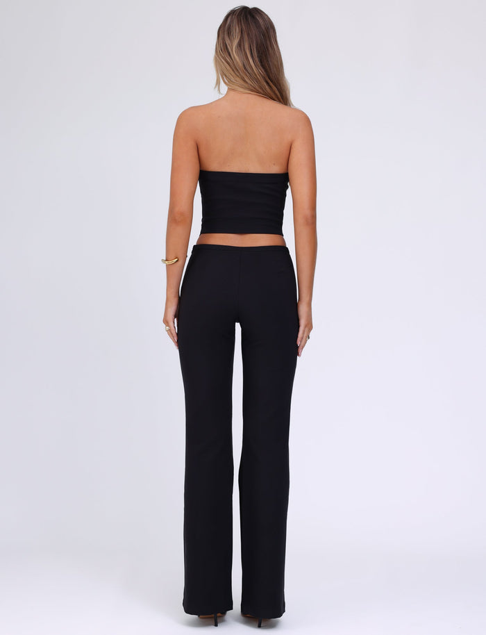 A woman with long, wavy hair stands facing away from the camera. She is wearing a black strapless top and TIGERMIST's AUDRIE PANT in black, made from suiting fabrication. A gold bracelet adorns her right wrist, and a faux leather belt cinches her waist. The background is plain white.