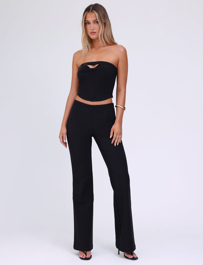 A woman with long, wavy hair wears the stylish AUDRIE TOP - BLACK from TIGERMIST, featuring a strapless neckline and a small front cutout. She pairs it with matching mid-length black pants and high-heeled shoes. Standing confidently against a plain white background, she accessorizes her look with a gold bracelet on her right wrist.