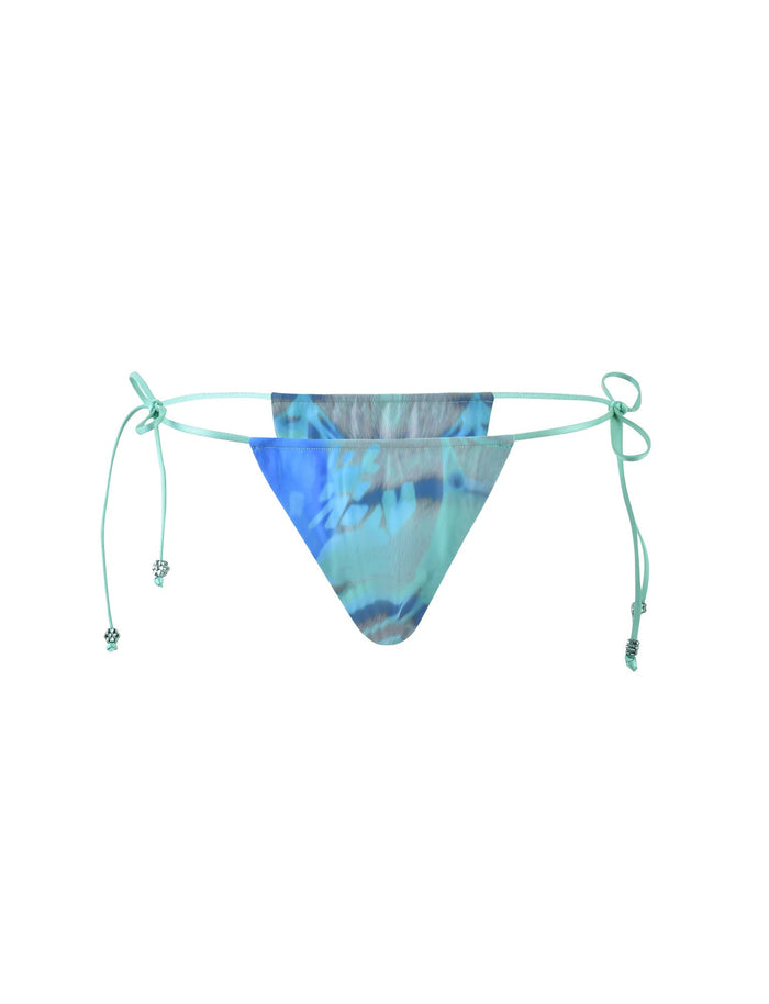 The image showcases the AVE Bikini Bottom from TIGERMIST, featuring a striking multi-colored tie-dye floral pattern. The light blue string ties, adorned with small decorative beads, offer an adjustable fit. The bikini bottom has a triangular front and matching side ties for customizable comfort.