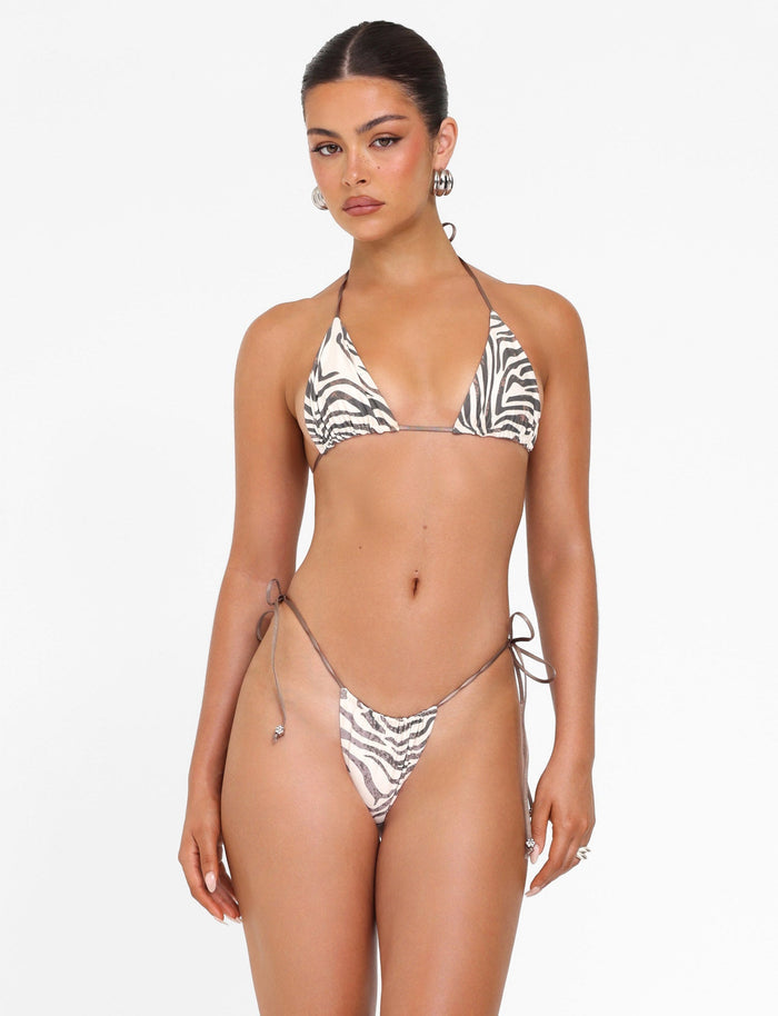 A woman models the TIGERMIST AVE BIKINI BOTTOM - NEUTRAL: ZEBRA, featuring adjustable side ties, against a plain white background. She accessorizes with hoop earrings and has her hair pulled back.