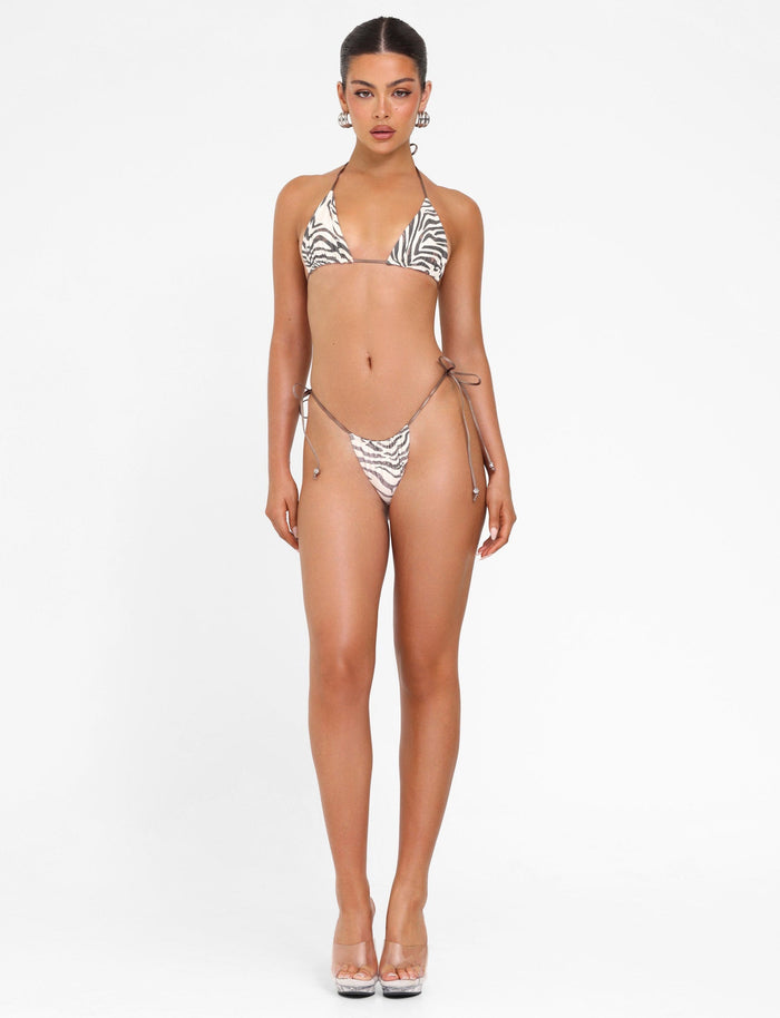 On a white background, a person confidently showcases the TIGERMIST AVE BIKINI BOTTOM - NEUTRAL : ZEBRA with adjustable side ties. They wear clear high heels and have their long hair neatly tied back, gazing directly into the camera.