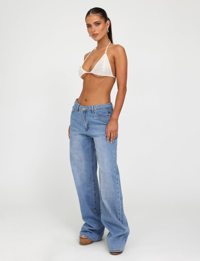A woman stands confidently against a plain white background, dressed in a white crocheted bikini top from Tiger Mist and the AVEREE JEAN - BLUE : MID WASH relaxed-fit jeans by TIGERMIST, which sit low on her hips. Her hair is pulled back, and she gazes towards the camera with a relaxed expression.