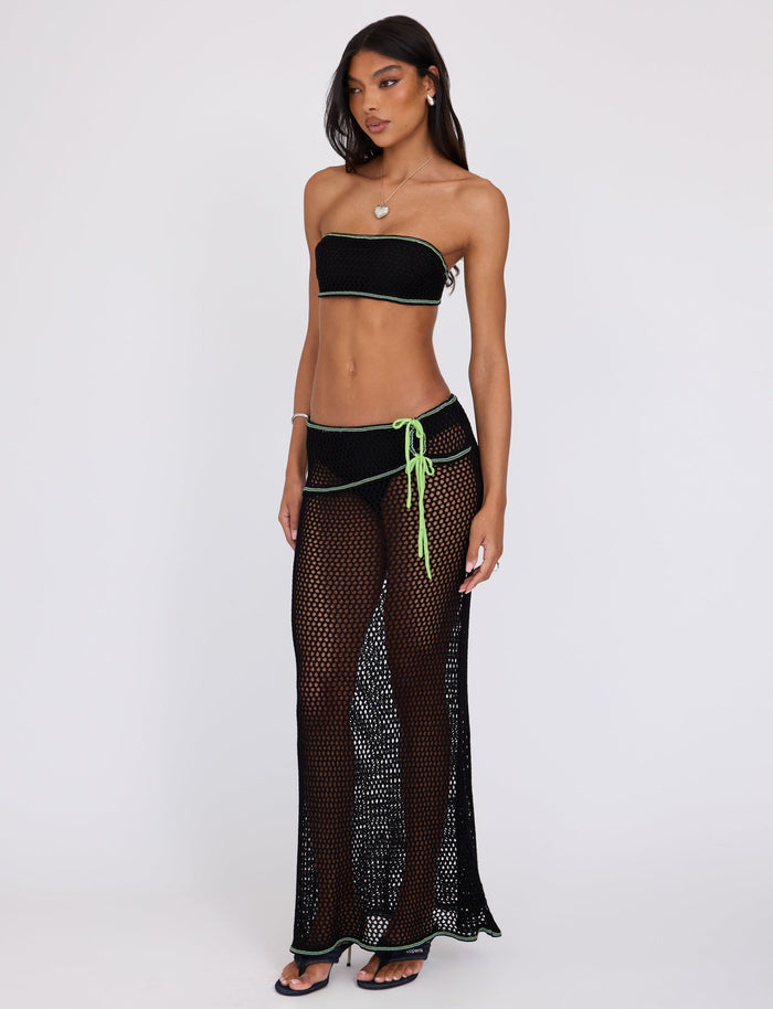 A woman stands against a plain background wearing a black bandeau bikini top and the TIGERMIST AZARIA SKIRT - BLACK, featuring a green waistband and drawstring. She accessorizes with a heart necklace, hoop earrings, and black sandals. Her long hair is worn loose.