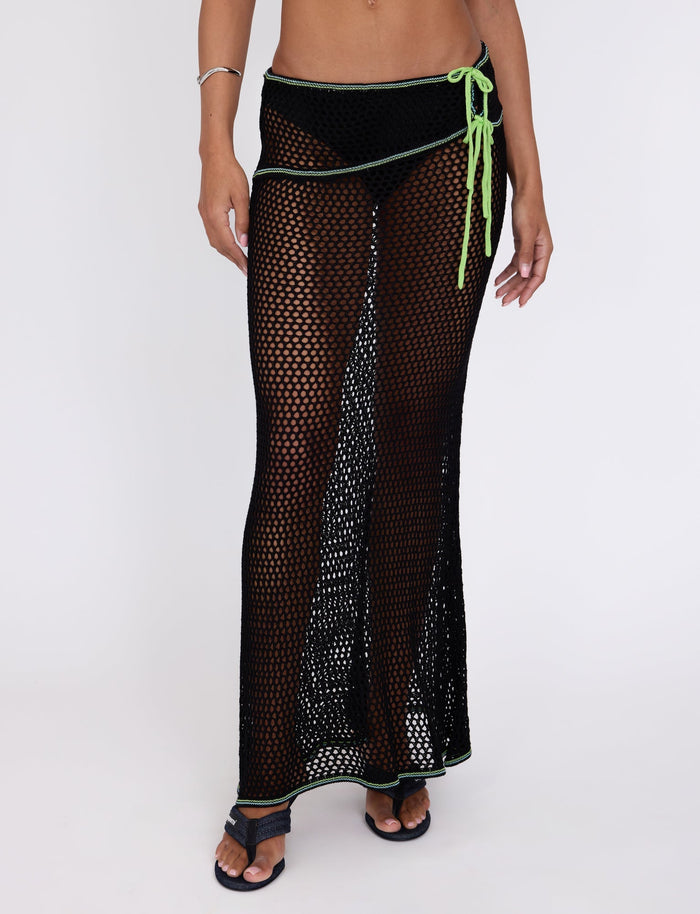 A person wearing the "Azaria Skirt - Black" from TIGERMIST, featuring green trim and a drawstring, is standing with their hand resting on their hip. The long, semi-transparent skirt reveals the swimsuit underneath. They are also wearing black sandals and a silver bracelet on one wrist.
