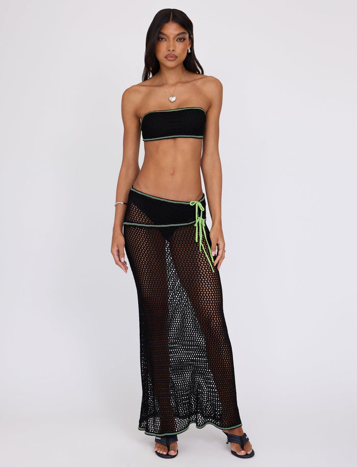 A woman stands modeling a TIGERMIST ensemble which includes the AZARIA SKIRT - BLACK, a long, sheer mesh piece worn over high-waisted bikini bottoms with a green string tie on the left hip. She pairs this with a strapless bandeau top and black sandals.