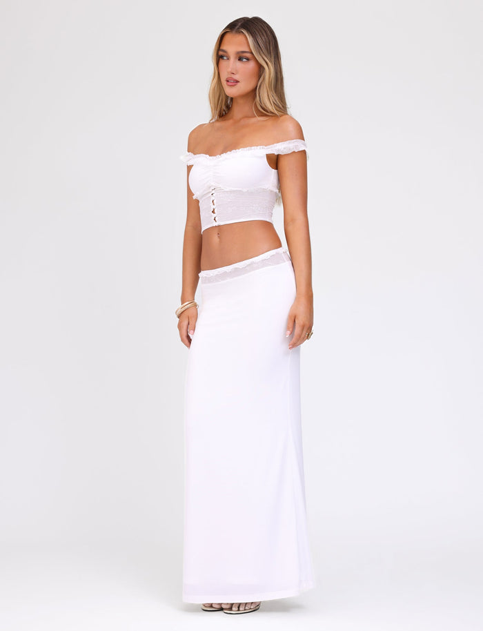A woman stands posing against a plain white background, wearing a white off-shoulder crop top with buttons down the front and the TIGERMIST BENTON SKIRT in white, which features a delicate lace waistband. She has long, wavy blonde hair and is accessorized with bracelets. She looks slightly to the side.