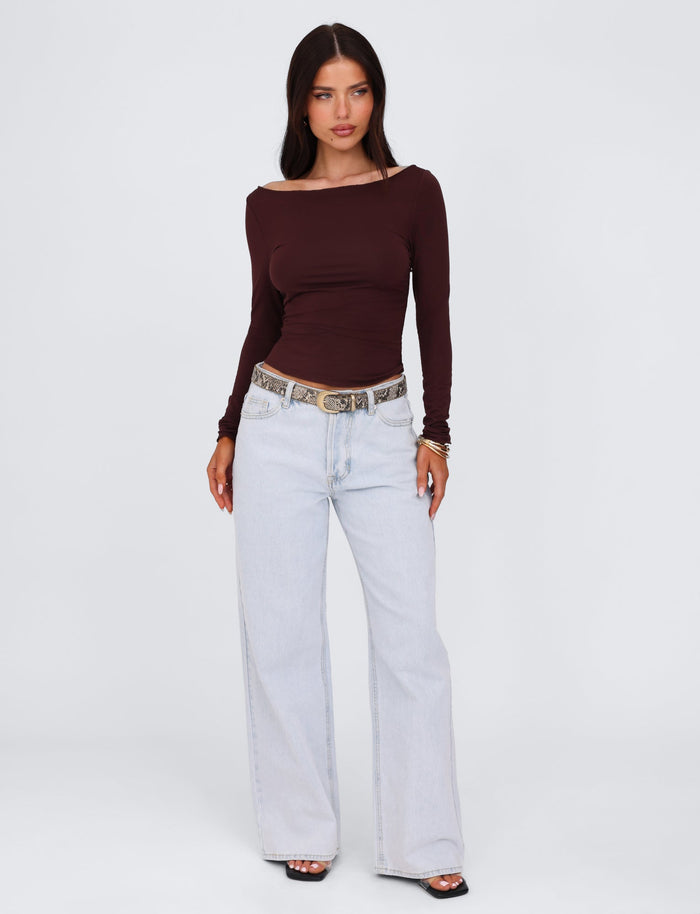 A woman with long dark hair stands against a plain background. She is wearing a chocolate brown Bianka top from TIGERMIST, styled in a bodycon silhouette, paired with light blue wide-leg jeans, a snakeskin patterned belt, and black open-toe shoes. She has a neutral expression and her hands are by her sides.