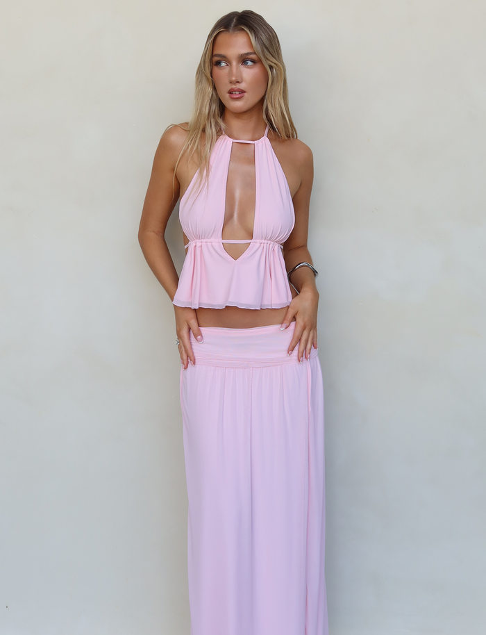 A woman with long blonde hair is wearing a baby pink, halter neck top with a triangle cutout and the matching TIGERMIST BIRDEE SKIRT in Baby Pink. She stands against a plain, light-colored wall, looking slightly to the right with her arms relaxed at her sides.