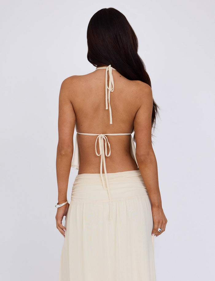 A woman with long dark hair stands with her back to the camera, wearing the stylish TIGERMIST BIRDEE TOP in white and a matching flowing skirt. Her back is mostly bare, except for the ties of the halter top. She appears to be in a well-lit studio with a plain white background.