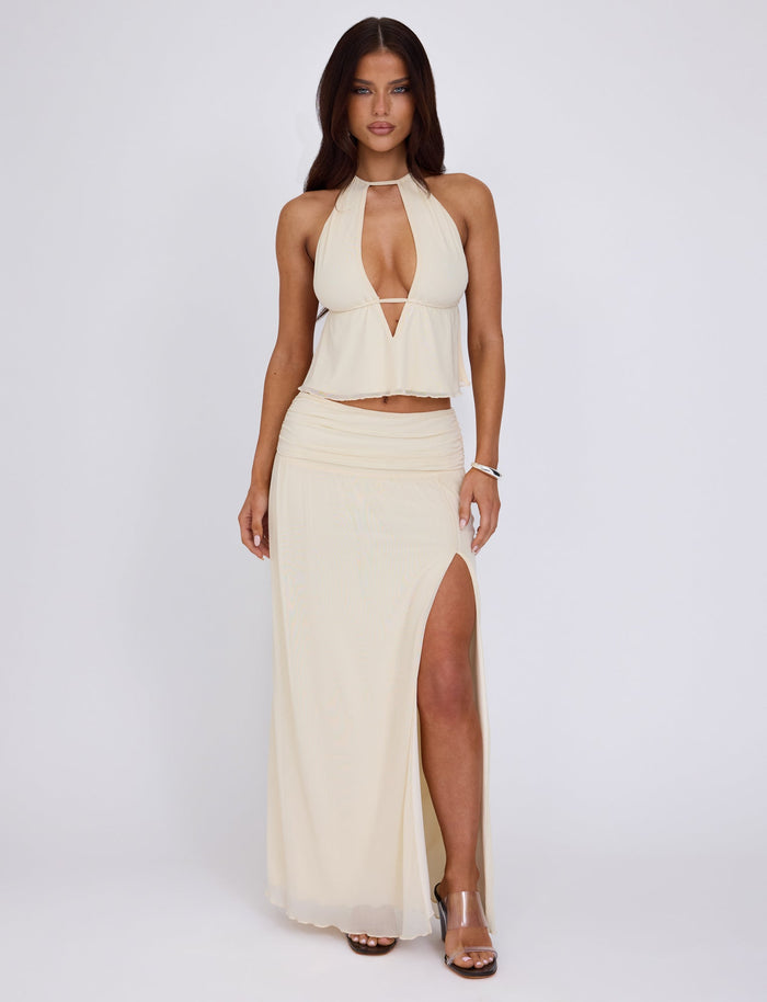 A woman with long brown hair is wearing the BIRDEE TOP in White: Cream with a plunging neckline and a matching long skirt with a high slit, both by TIGERMIST. She stands against a plain white background, one hand by her side and the other resting on her hip. She is wearing clear high-heeled shoes.