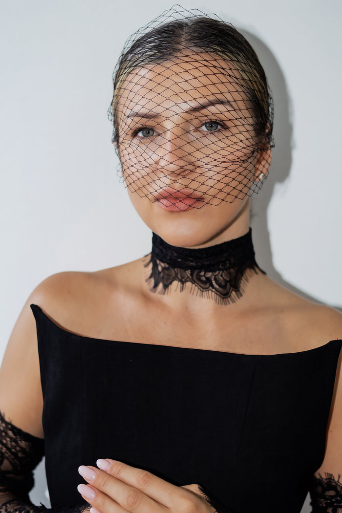 A woman wearing a black lace choker necklace with a matching black birdcage veil on her head, paired with a black off-the-shoulder top. She is posed against a plain light gray background.