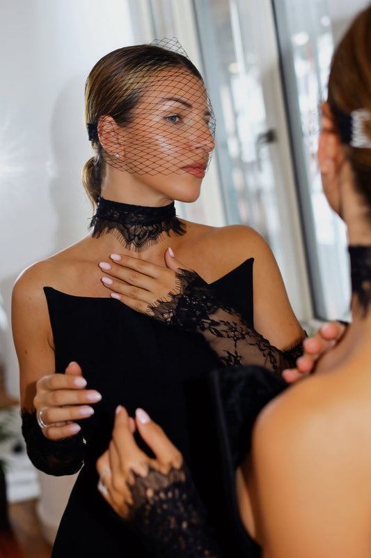 A woman wearing a black lace choker, matching lace gloves, and a black veil with a feather headpiece, seen in a mirror reflection while adjusting her dress.