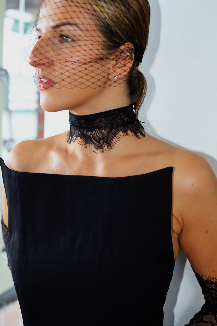 A woman wearing a black lace choker collar with attached fishnet veil and a black off-the-shoulder dress, shown in a close-up portrait against a light indoor background.