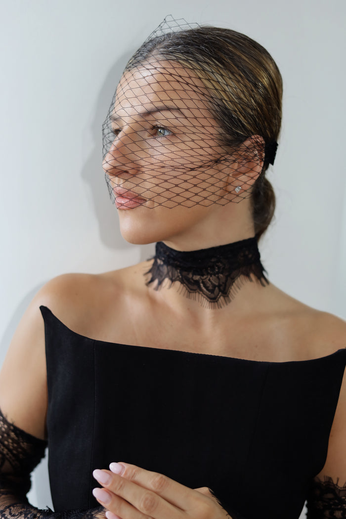 A woman wearing a black lace choker and a black bird's-eye net veil over her face, paired with a black off-the-shoulder top.