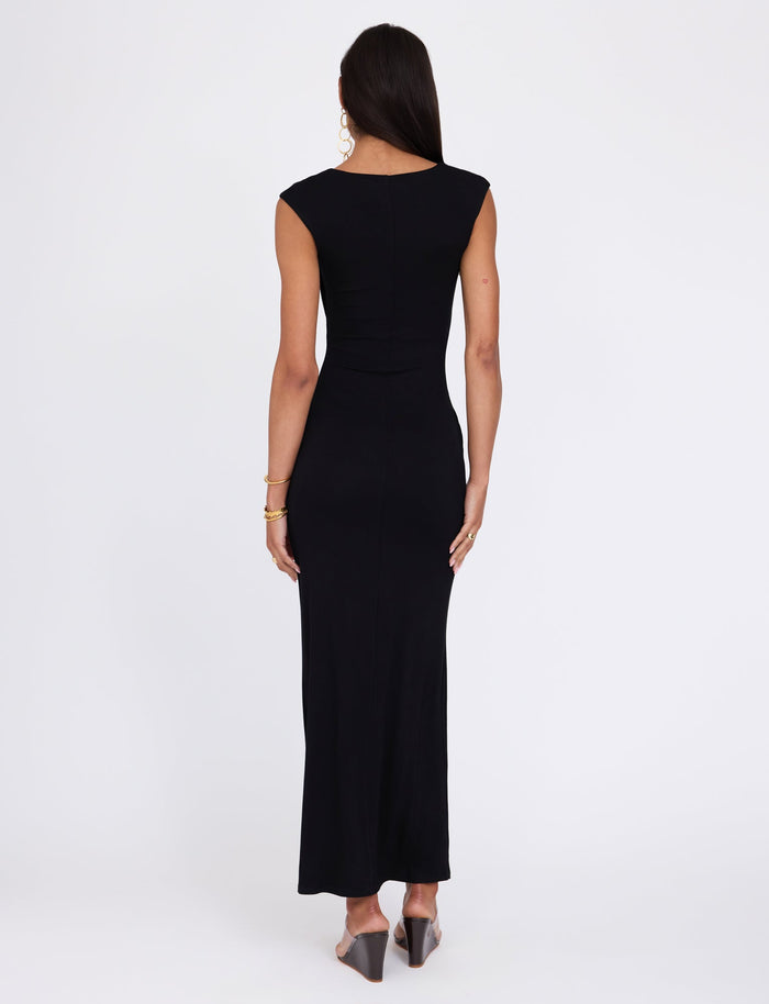 A woman with long brown hair is standing with her back to the camera, wearing the fitted, sleeveless BLAKELY DRESS - BLACK by TIGERMIST that reaches her ankles. She is accessorized with gold hoop earrings, gold bracelets, and black heeled mules. The background is plain white.