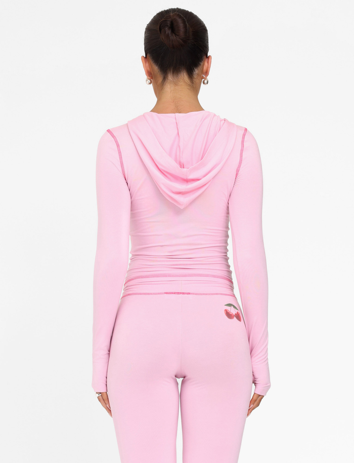 A woman with her dark hair in a bun wears the TIGERMIST BLAYZE HOODIE - PINK: SOFT PINK, paired with matching leggings featuring a small cherry detail at the waistband. She faces away against a plain white background.