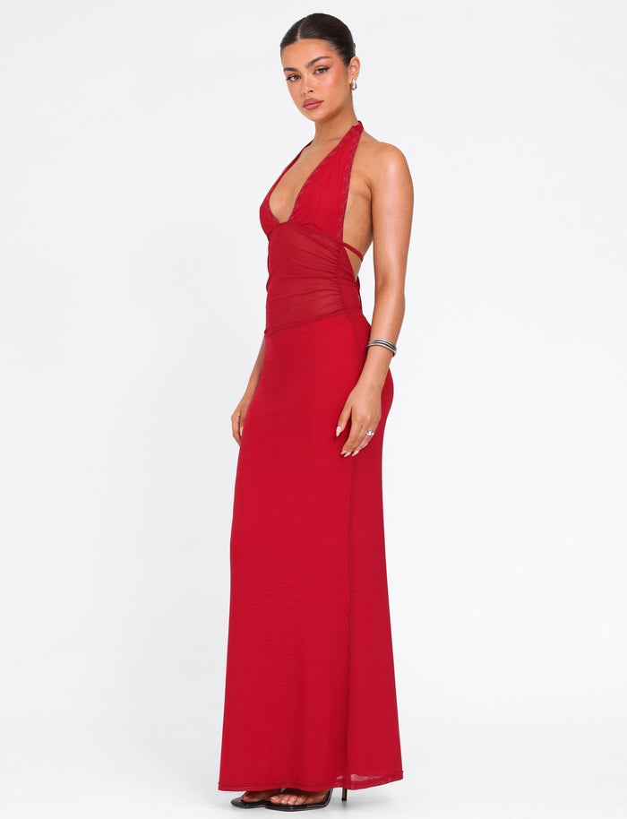 A woman stands in profile against a white background wearing the TIGERMIST BRESILY MAXI DRESS in red, a sleeveless floor-length gown with halter straps and a fitted bodice, styled with black heels and minimal jewelry. Her hair is pulled back.