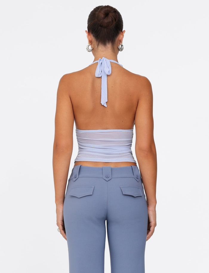 From behind, a person with their hair styled in a bun is dressed in the Tigermist Bresily Top in Steel Blue, complemented by high-waisted pants. The pants, highlighted by pocketed buttons, create a striking contrast against the simple white backdrop.